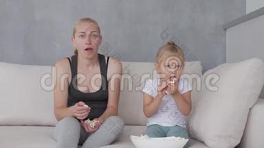 一个女人和一个小女孩坐在柔软的沙发上，吃着爆米花，看着电视屏幕。